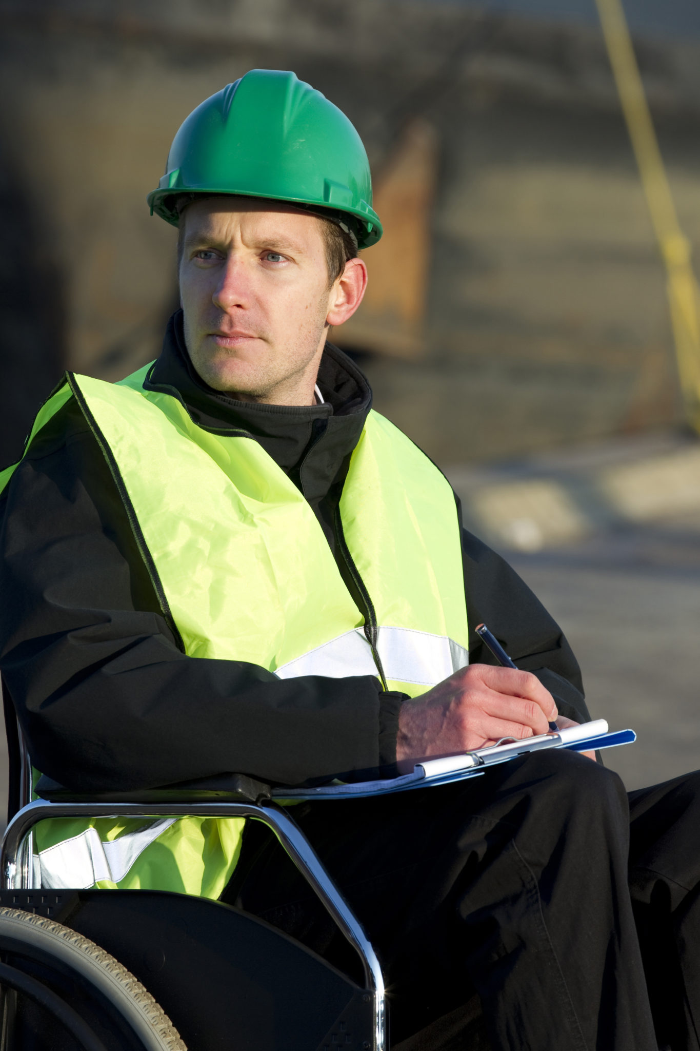 Construction supervisor in wheelchair, wearing safety vest and writing on notepad at construction site